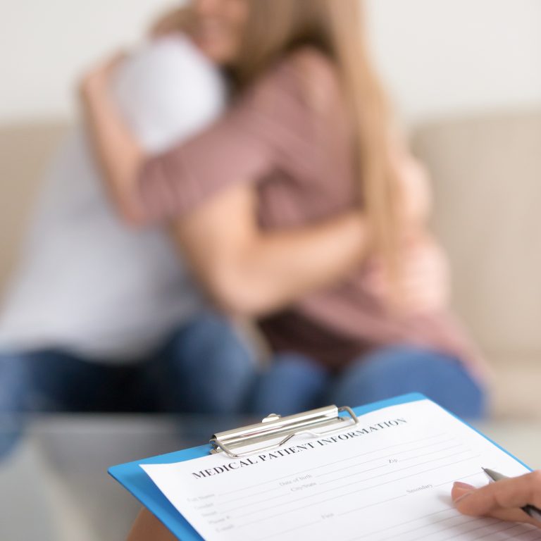 Close up of female psychotherapist holding clipboard, filling information about patients while couple embracing on couch in the background, doctor making notes in medical card in psychologists office